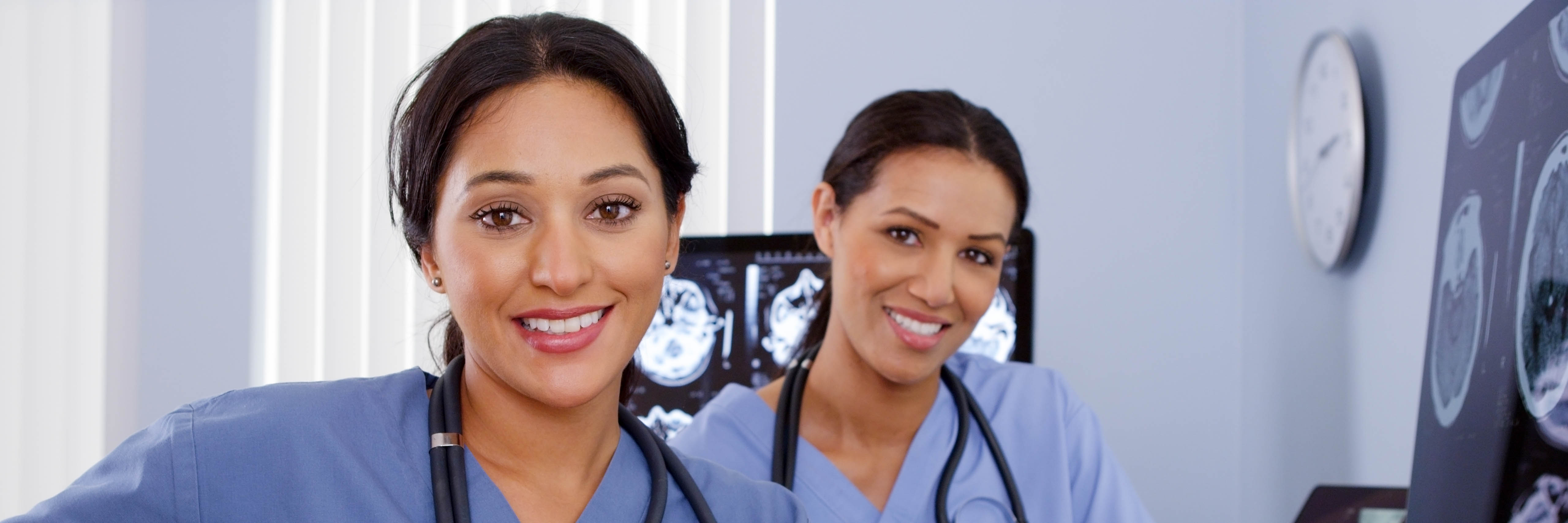 Two young women clinicians smiling, with stethoscopes around their necks, sitting at their work stations in front of brain scan x-ray images.
